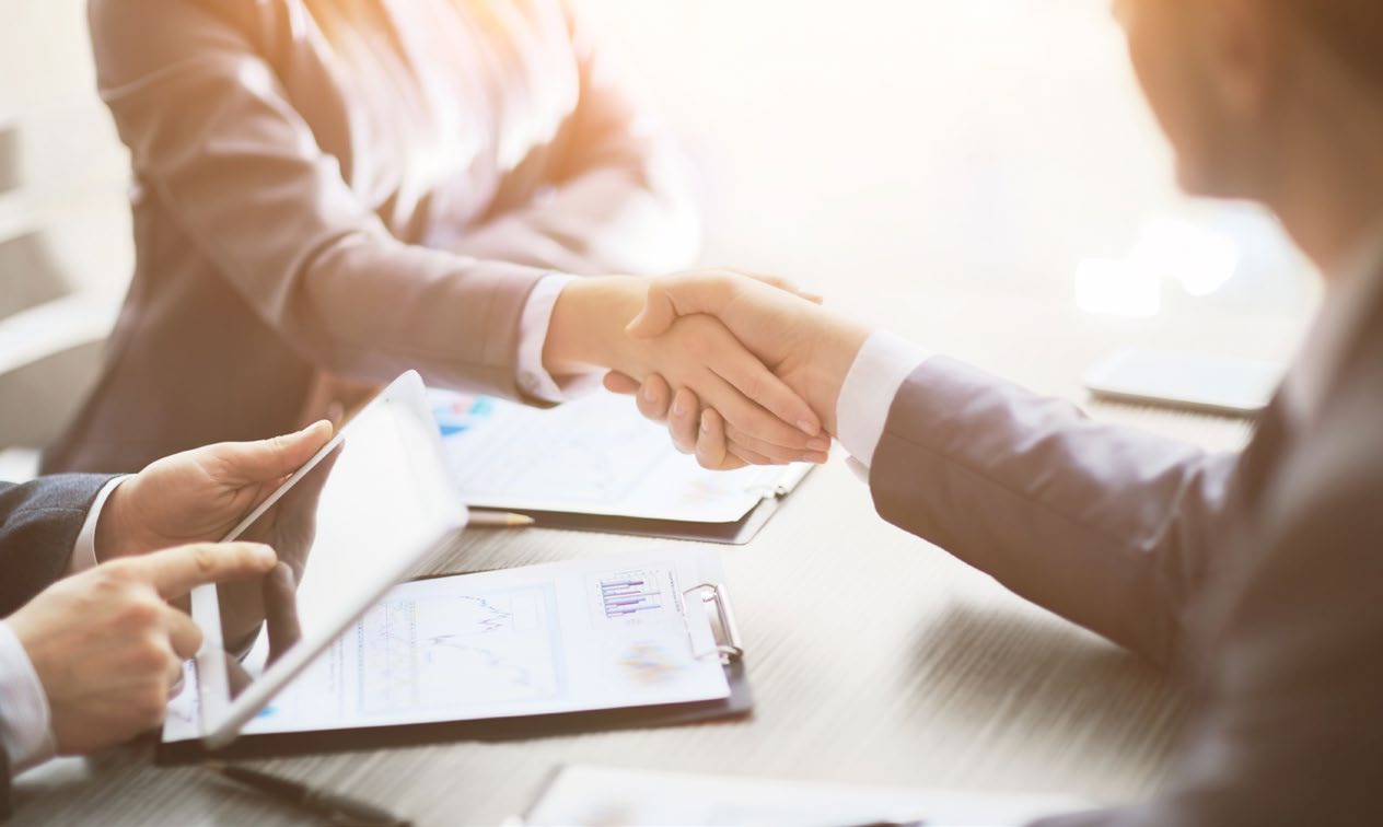 Two people shaking hands across a table spread with printed documents