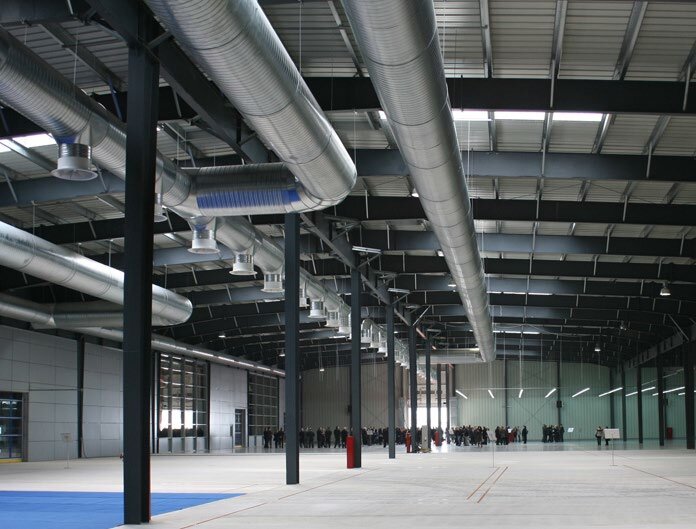 Un grand hall industriel vide aux colonnes d’acier apparentes, avec des gaines de ventilation circulaires au plafond