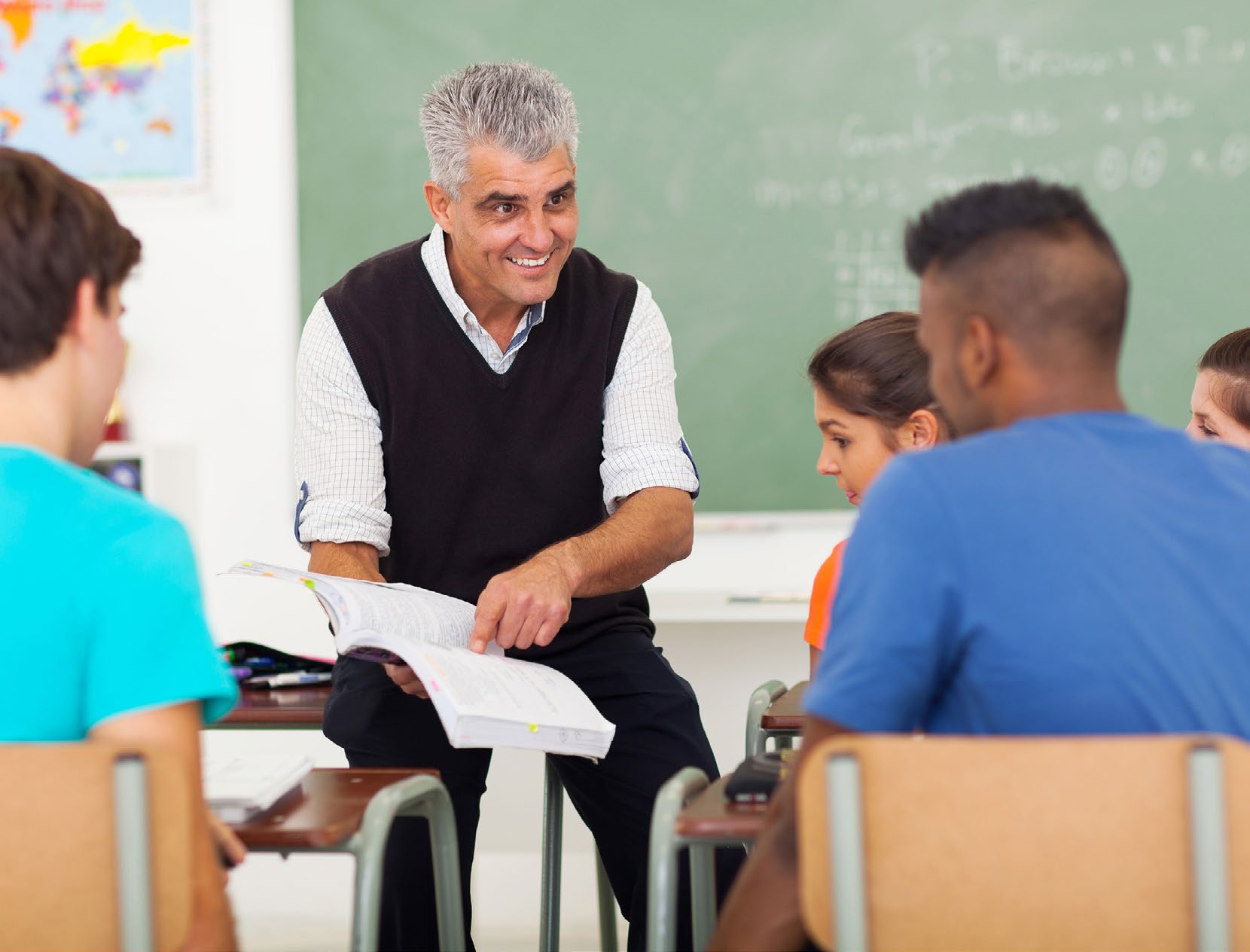 A teacher sitting with an open book, talking with students in a classroom