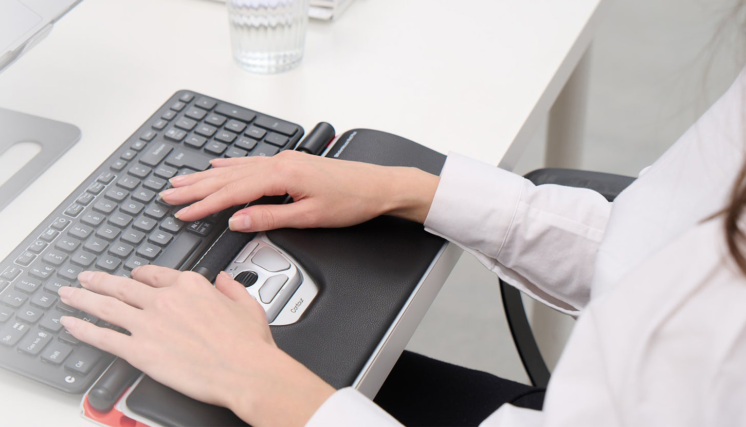 Hände an einer Contour RollerMouse und einer Tastatur an einem weißen Schreibtisch
