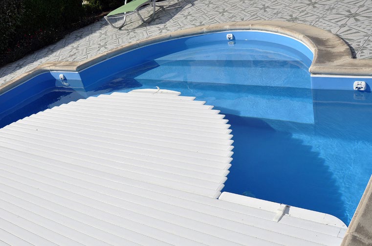 An automatic slatted pool cover partly rolled across a blue swimming pool
