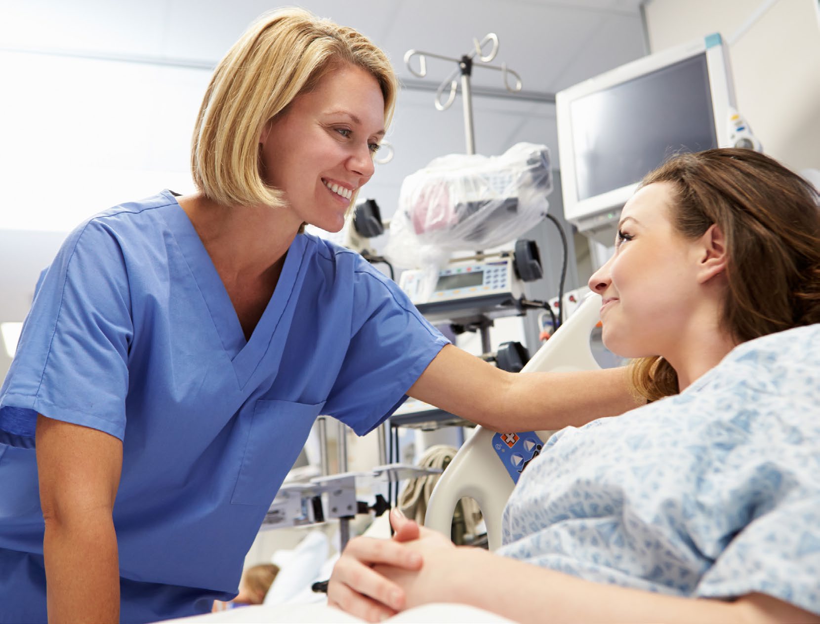 A nurse in scrubs leaning in to talk with a patient in a hospital bed