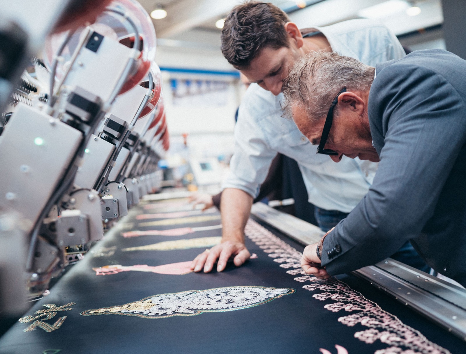 Dos compañeros inclinados sobre una máquina de bordar revisando un panel terminado