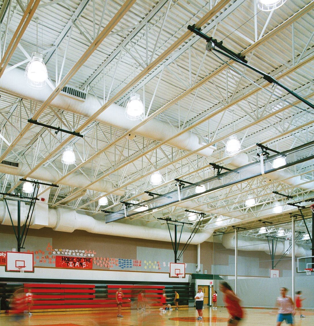 Un gymnase scolaire avec des fermes de toit en acier blanc et de longues gaines de ventilation textiles au-dessus d’un terrain de basket