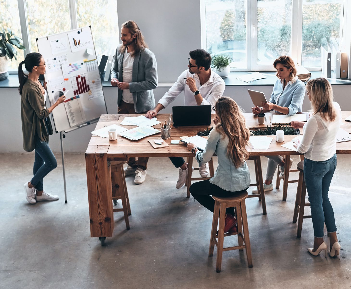 A team gathered around a long wooden table, one person presenting at a whiteboard of charts