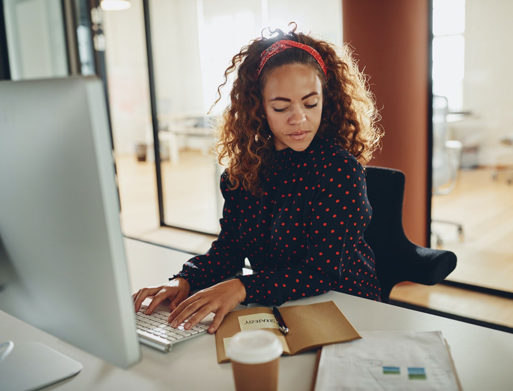 A woman working at a desktop computer beside a stack of paper documents