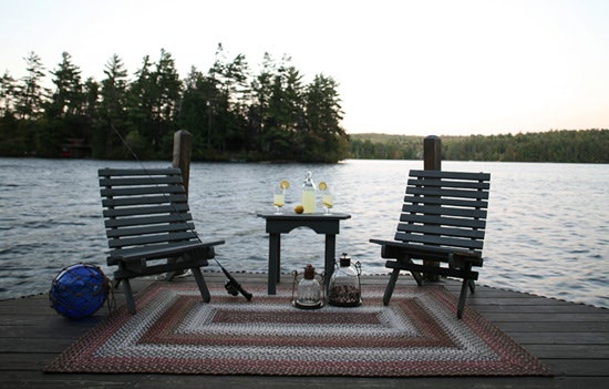 Deux fauteuils en bois et une petite table sur un ponton au bord d’un lac, posés sur un tapis tressé