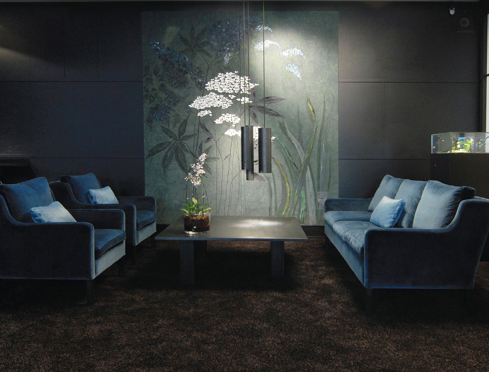 A dark, softly lit showroom lounge with blue velvet sofas and a floral mural